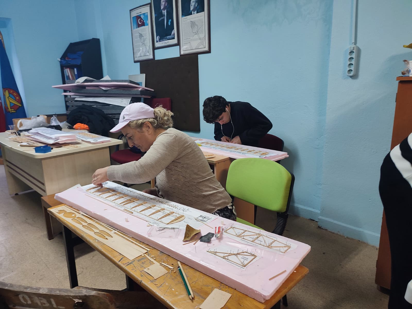 İki öğrenci tamamlanmakta olan bir model uçağın önünde duruyor. Uçak, öğrencilerin emeğinin bir sonucu olarak atölyenin başarısını simgeliyor.